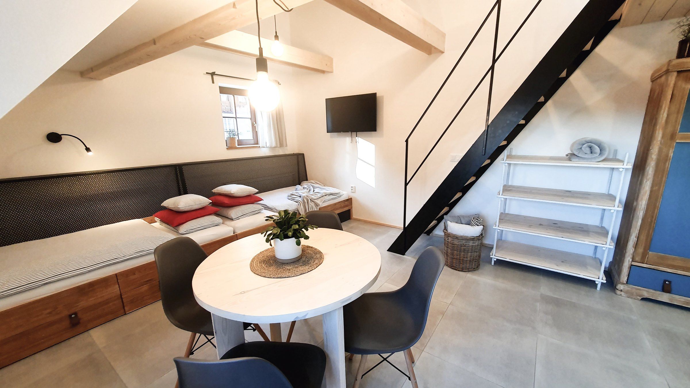 Apartmán III: lůžka za sebou s čalouněným opěradlem, kulatý stůl, černé schody na galerii a modrá skříň
