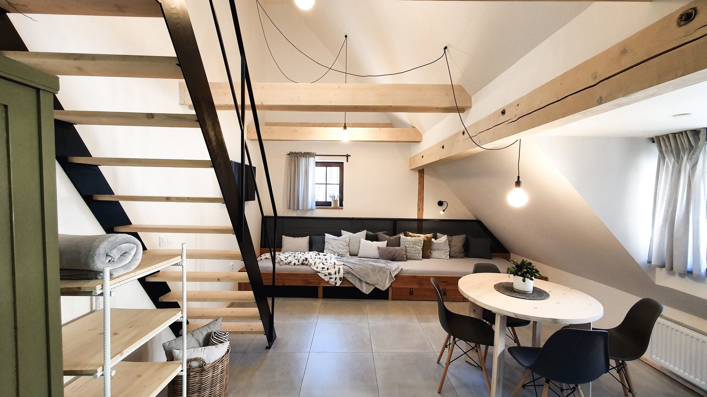 Apartmán IV: schody na galerii, lůžko s polštáři u okna a kulatý stůl s židlemi